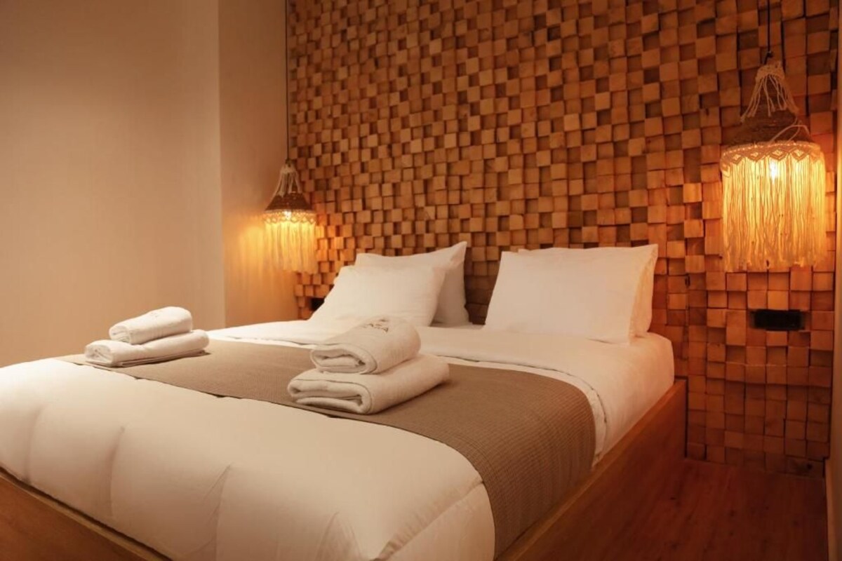 A neatly made bed with white linens and towels, a wooden textured wall behind, and two wall-mounted lamps.