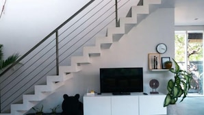 A modern living room with a staircase, a flat-screen TV, a white entertainment unit, and a potted plant.