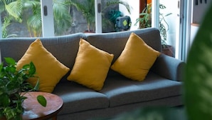 A grey sofa with three yellow cushions, a small round wooden table with a plant, and a view of green foliage through a glass door.
