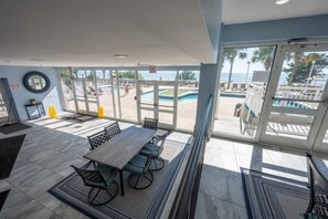 A spacious indoor area with a large table and chairs, large windows offering a view of a pool and beach.
