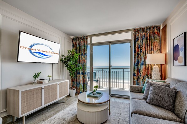 A modern living room with a television displaying 'Ocean Wave Rentals,' a large potted plant, a round coffee table, a sofa, and a balcony with a view of the ocean.