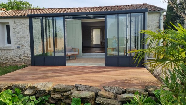 A modern house with a stone exterior, a wooden deck, and a glass door leading to a room with a bench and a chair.