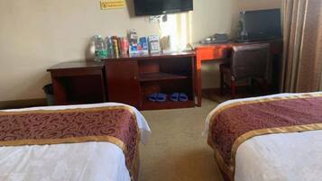 A hotel room with two beds, a desk, a chair, a television, and a shelf with various items.