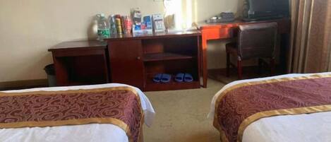 Habitación de hotel con dos camas, un escritorio, una silla, un televisor y una estantería con diversos objetos.