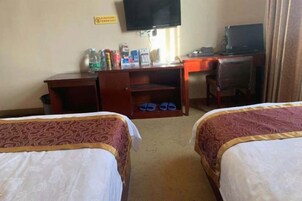A hotel room with two beds, a desk, a chair, a television, and a shelf with various items.