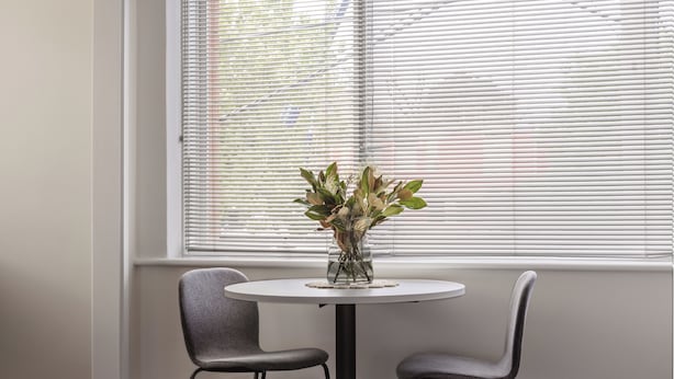 A small round table with two chairs, a vase with green plants, and a window with blinds. - MAC St Kilda by Melbourne Apartment Collection (St Kilda)