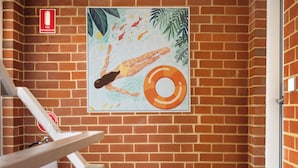 A hallway with a brick wall, a fire extinguisher, and a poster of a person diving with a ring. - MAC St Kilda by Melbourne Apartment Collection (St Kilda)
