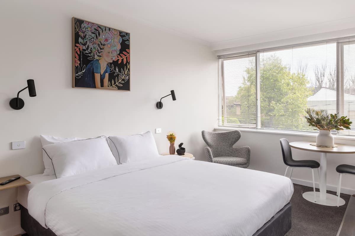 A hotel room with a large bed, a small table with two chairs, a potted plant, a painting on the wall, and a window with blinds.