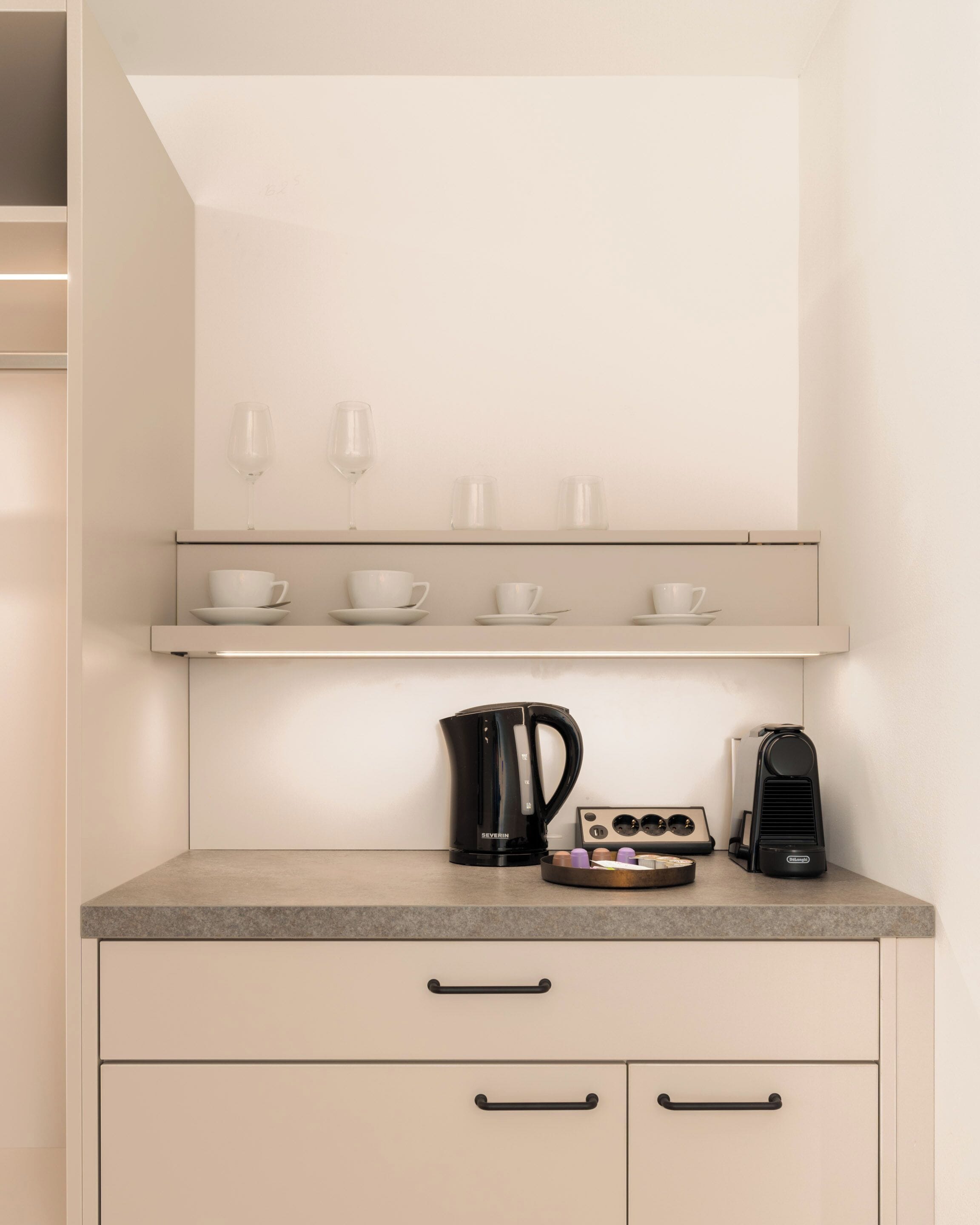Een kitchenette met een aanrecht, een koffiezetapparaat, een waterkoker en een koffiemachine. Op de plank erboven staan bekers en glazen.