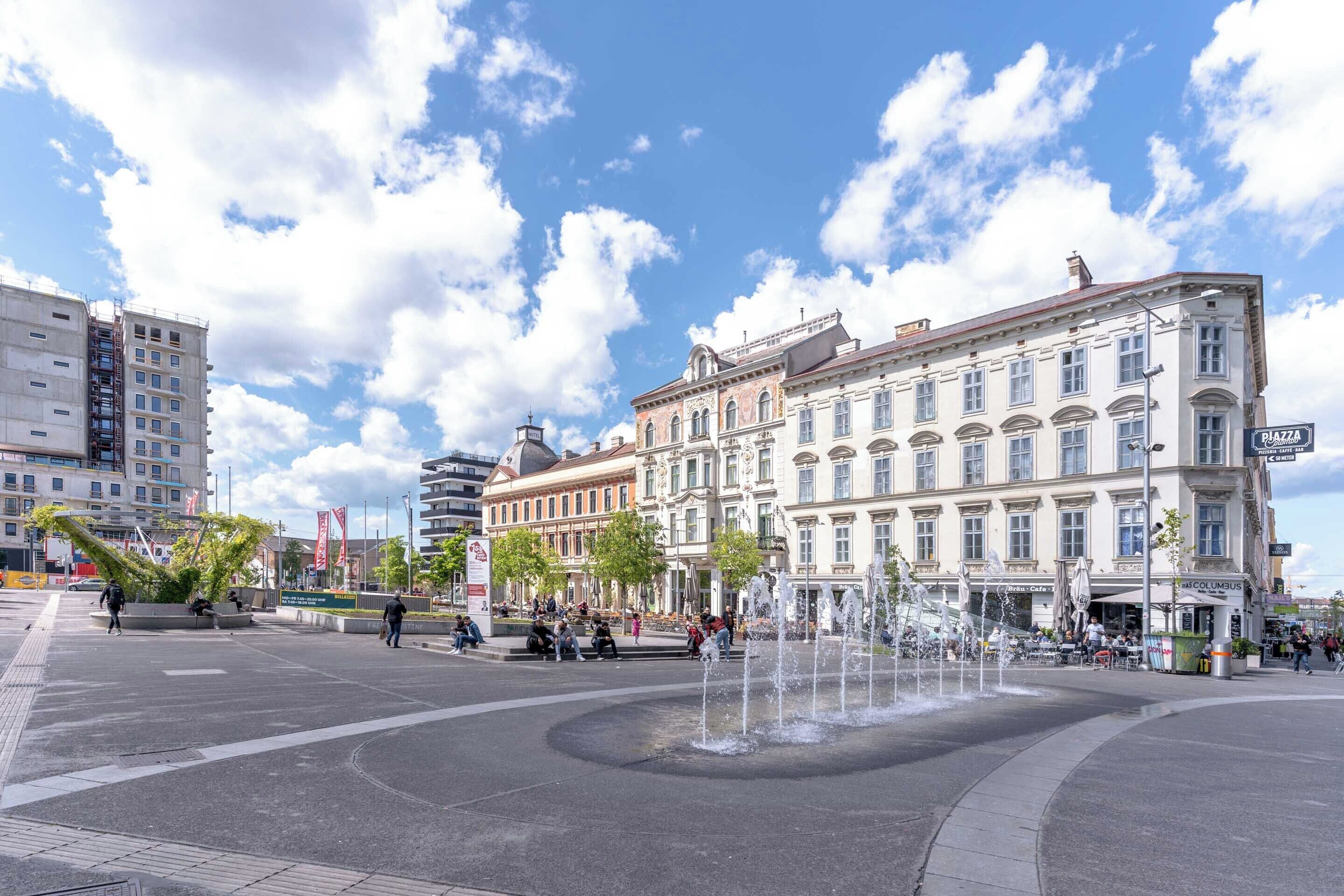 Une place publique avec une fontaine, entourée de bâtiments historiques et d’un immeuble moderne de grande hauteur.