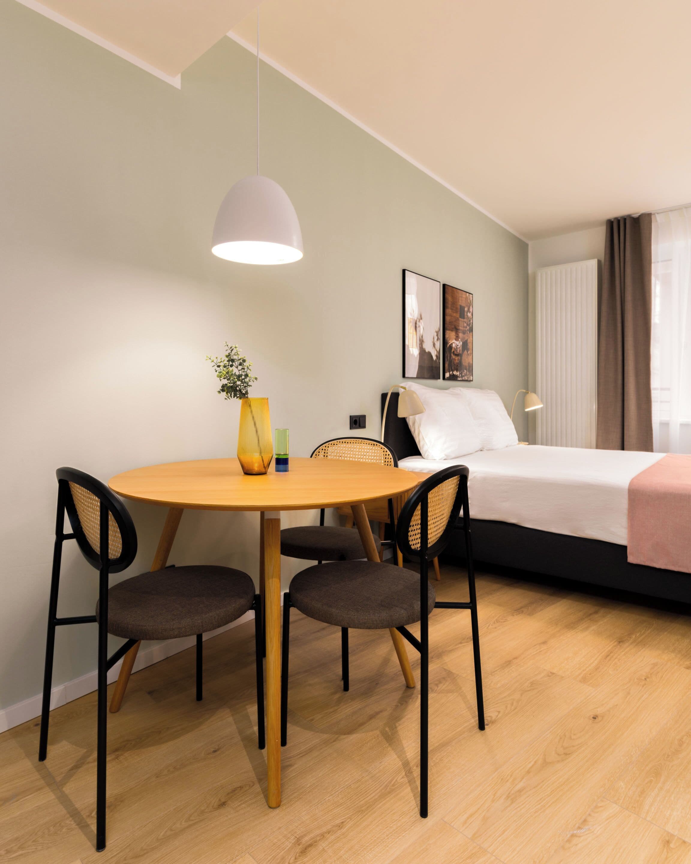 A modern hotel room with a wooden dining table, four chairs, a bed with a white mattress and a pink blanket, a bedside lamp, and a large window with curtains.