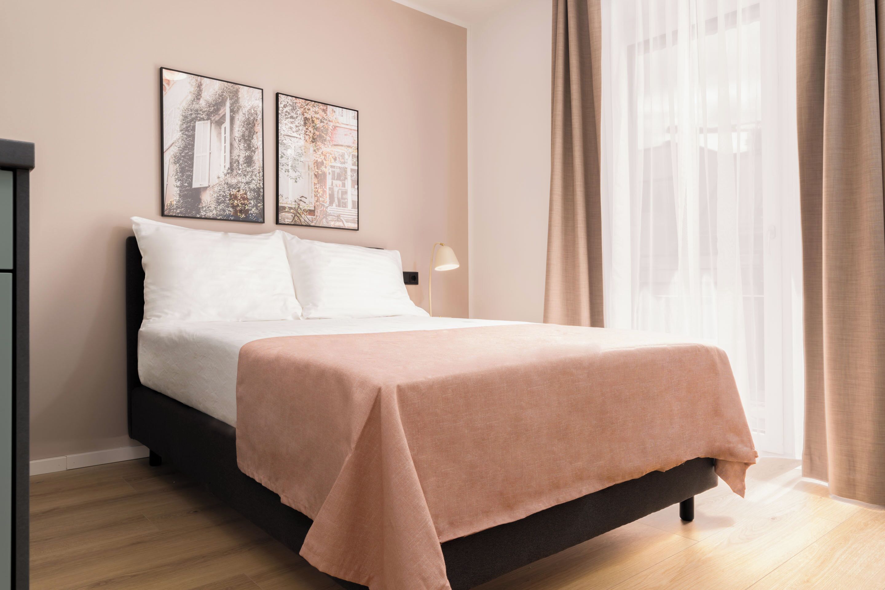 A neatly made bed with a white and peach-colored bedspread, two framed pictures on the wall, a bedside lamp, and sheer curtains.