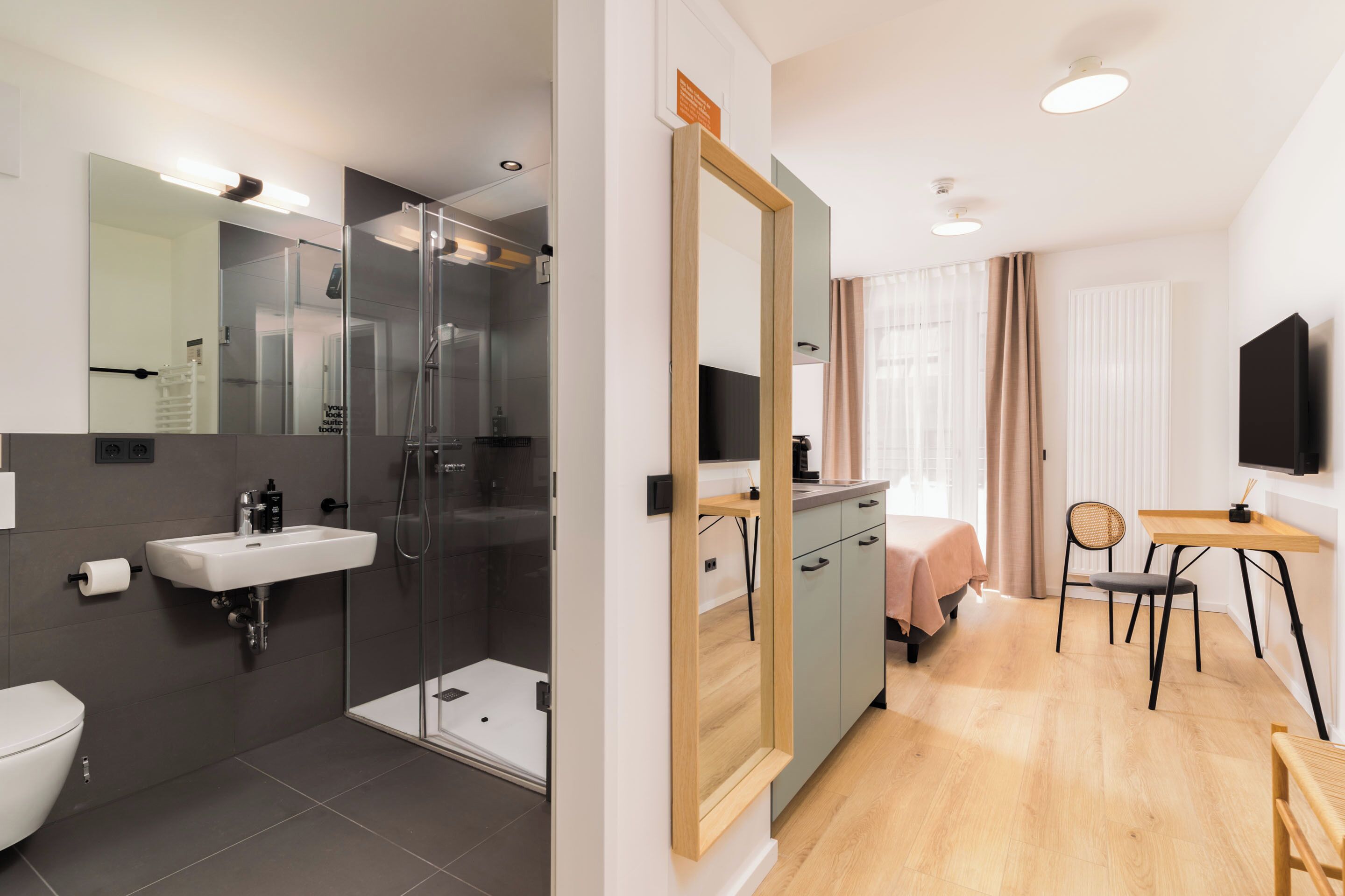 A modern bathroom with a shower, sink, and toilet. There is a bed with a wooden headboard and a desk with a chair.