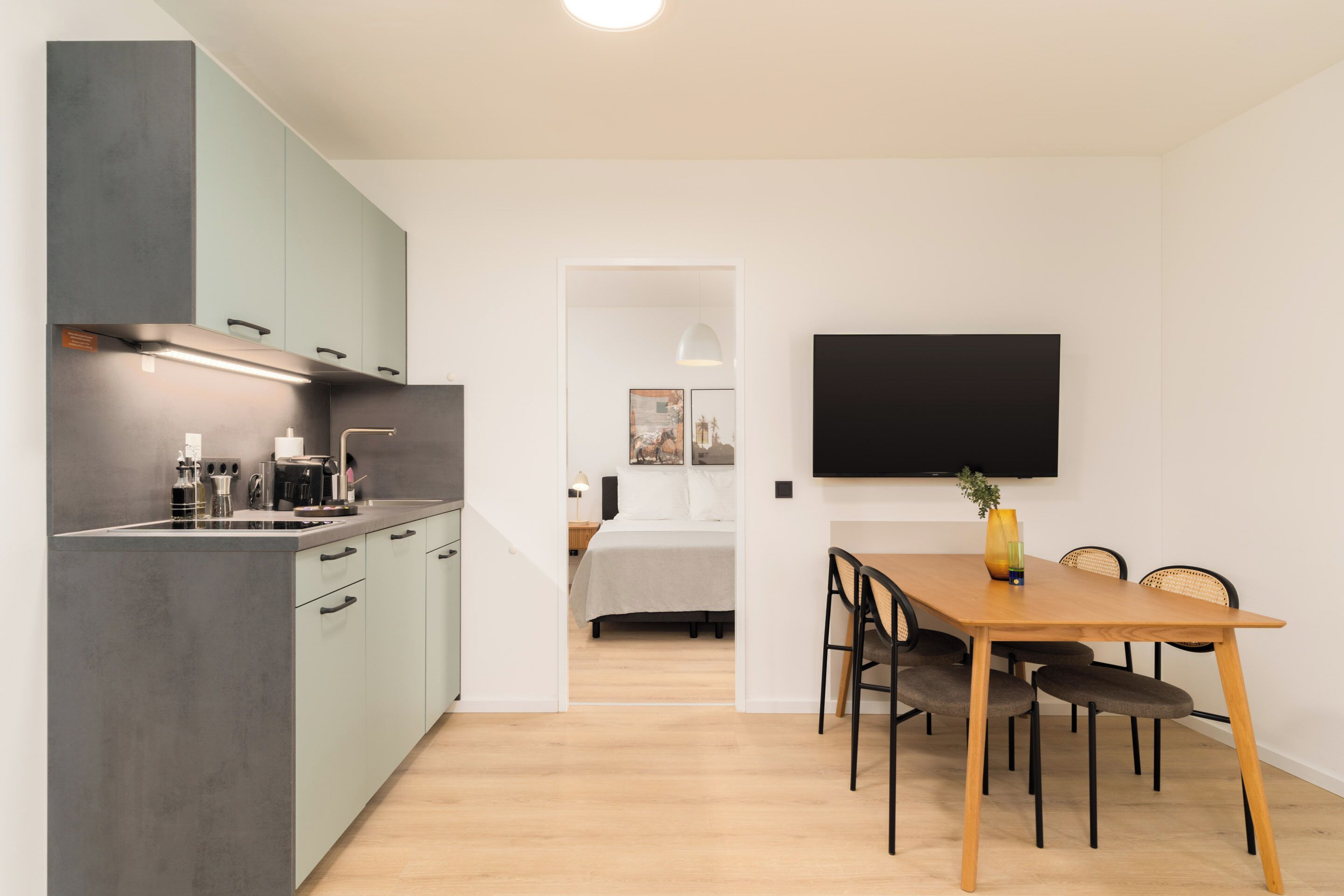 A modern kitchen with grey cabinets, a wooden dining table, and a flat-screen TV mounted on the wall.