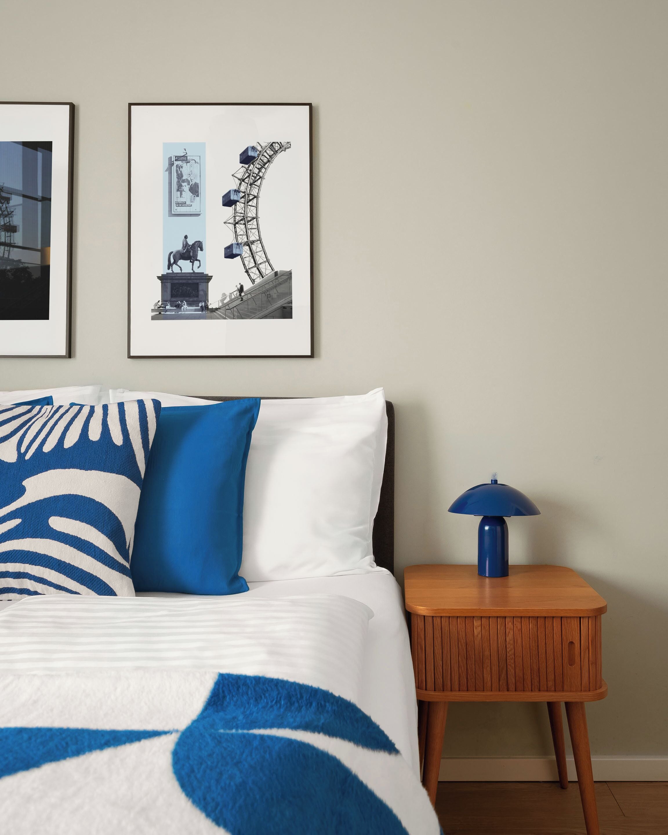 Een slaapkamer met een bed, kussens, een nachtkastje, een lamp en twee fotolijstjes aan de muur.