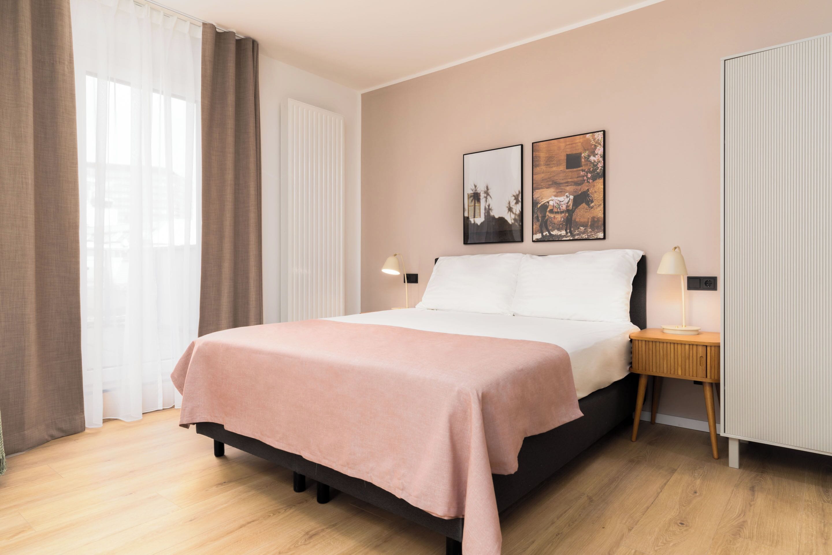 A neatly made bed with a pink bedspread, a bedside table with a lamp, and two framed pictures on the wall.