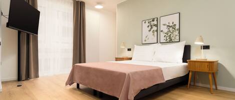 A bedroom with a bed, a flat-screen TV mounted on the wall, two bedside tables with lamps, and a window with curtains.