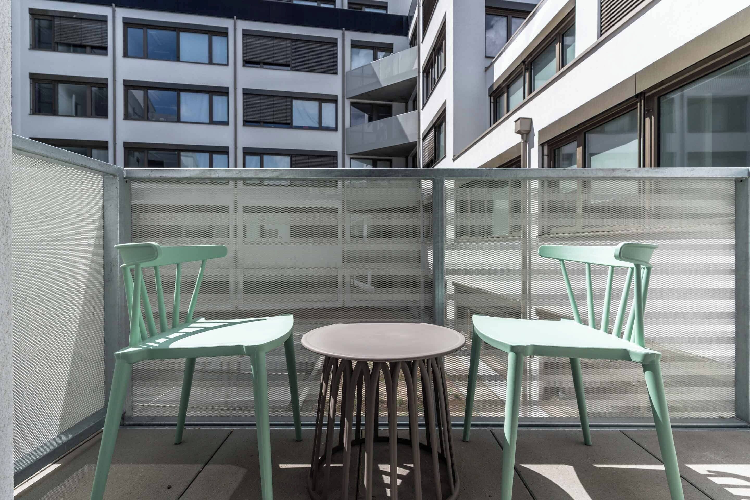 Un balcón con dos sillas y una mesa, con vista a un edificio moderno con múltiples ventanas.