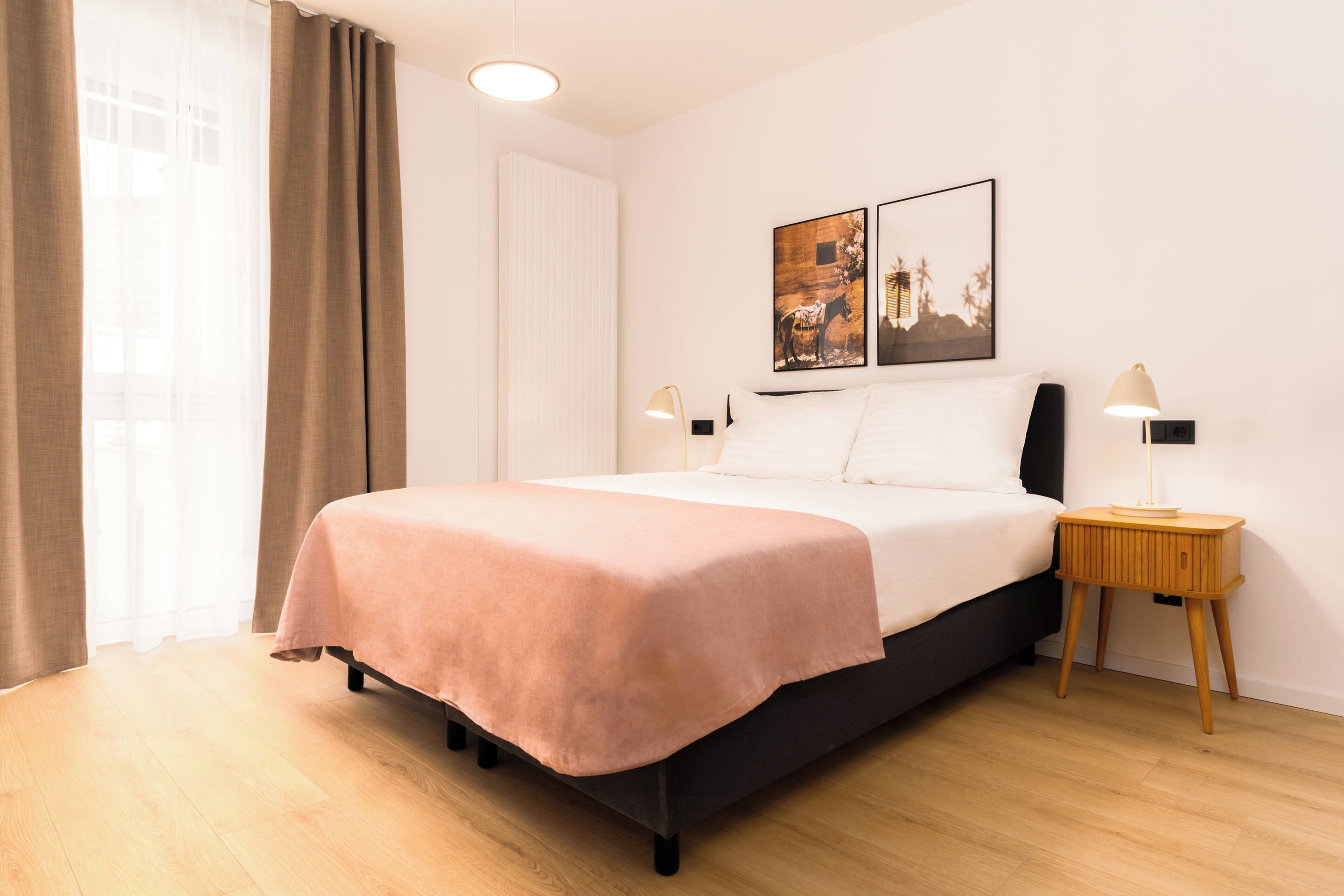 A neatly made bed with a pink bedspread, a bedside table with a lamp, and two framed pictures on the wall.