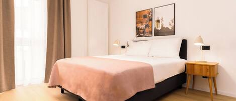 Una cama bien hecha con colcha rosa, una mesita de noche con lámpara y dos cuadros en la pared.