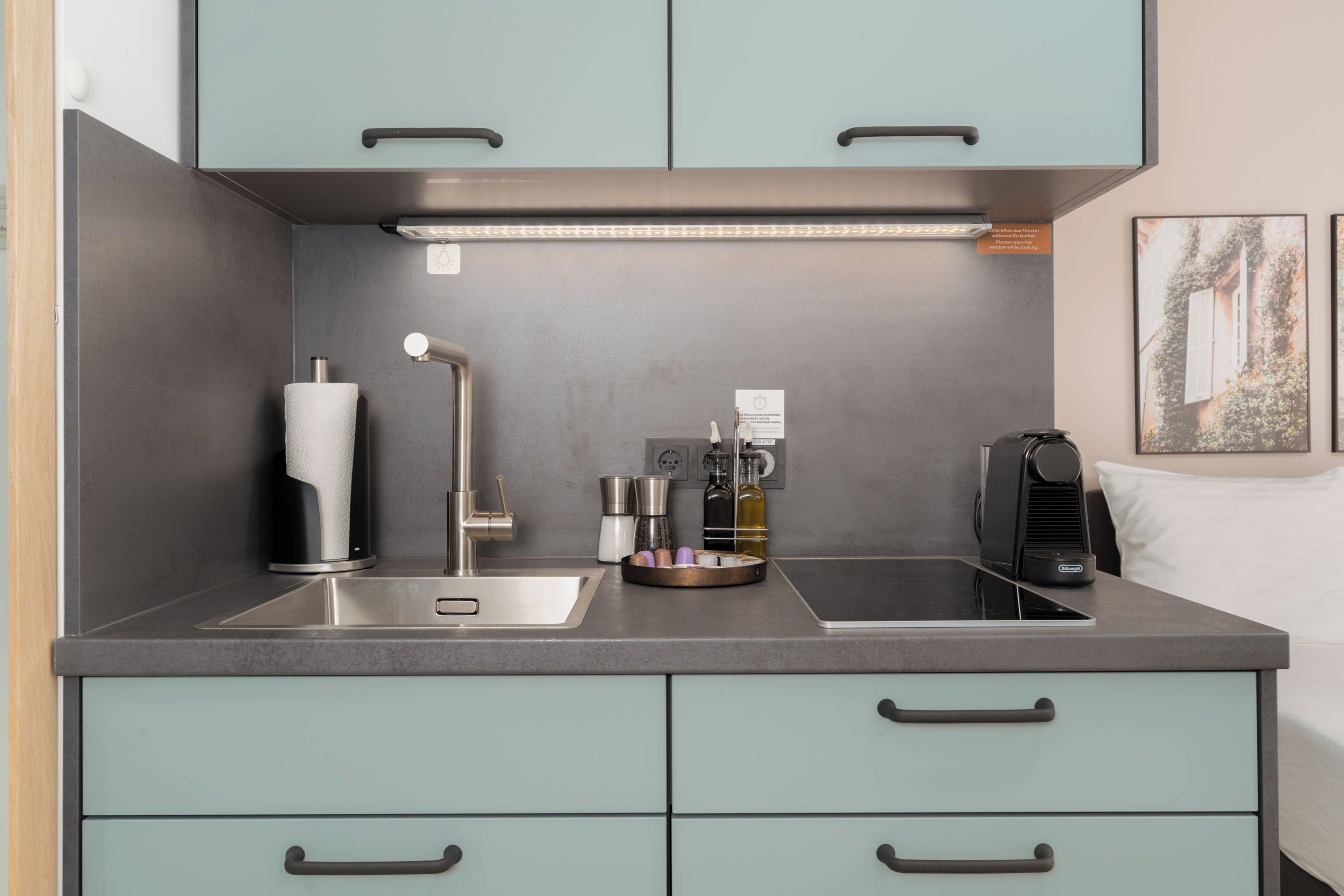A modern kitchenette with a stainless steel sink, a coffee machine, and a kettle. There are light blue cabinets and a countertop.