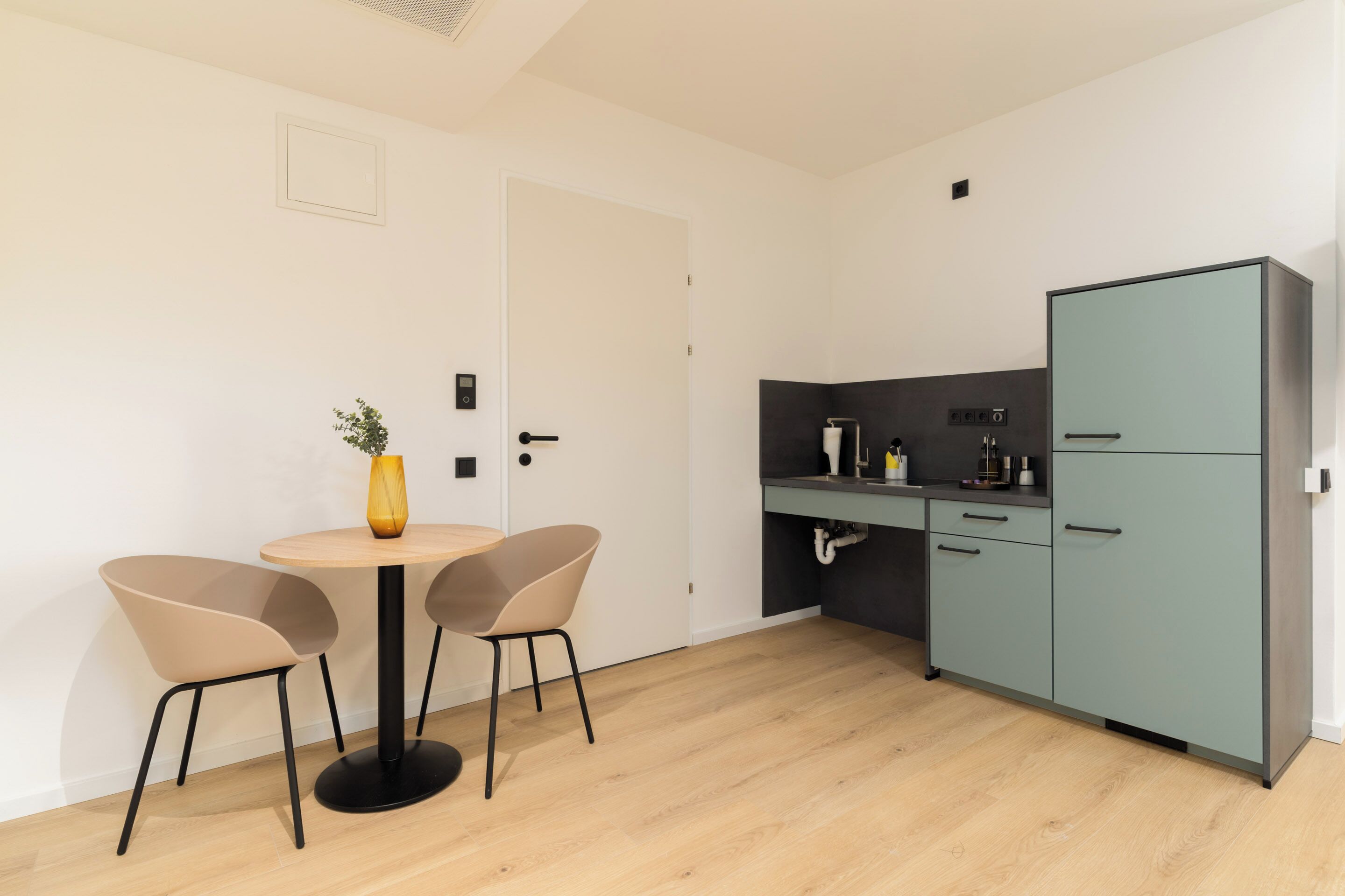 A compact kitchen with a round table and two chairs, a small cabinet, and a door.
