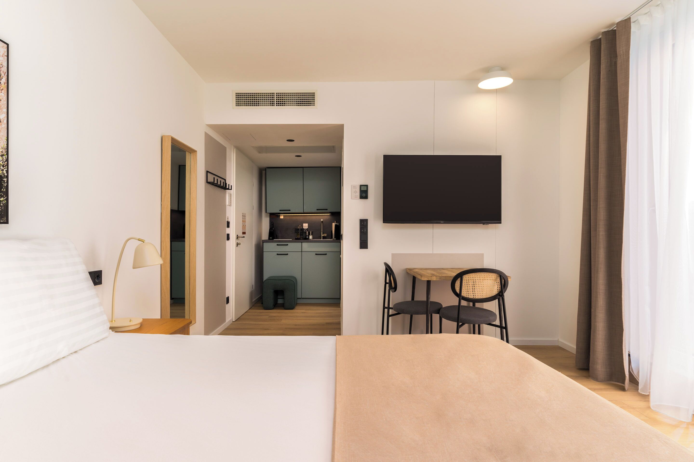 Une chambre d’hôtel avec un lit, un bureau, une chaise, une télévision et, au fond, une petite cuisine.