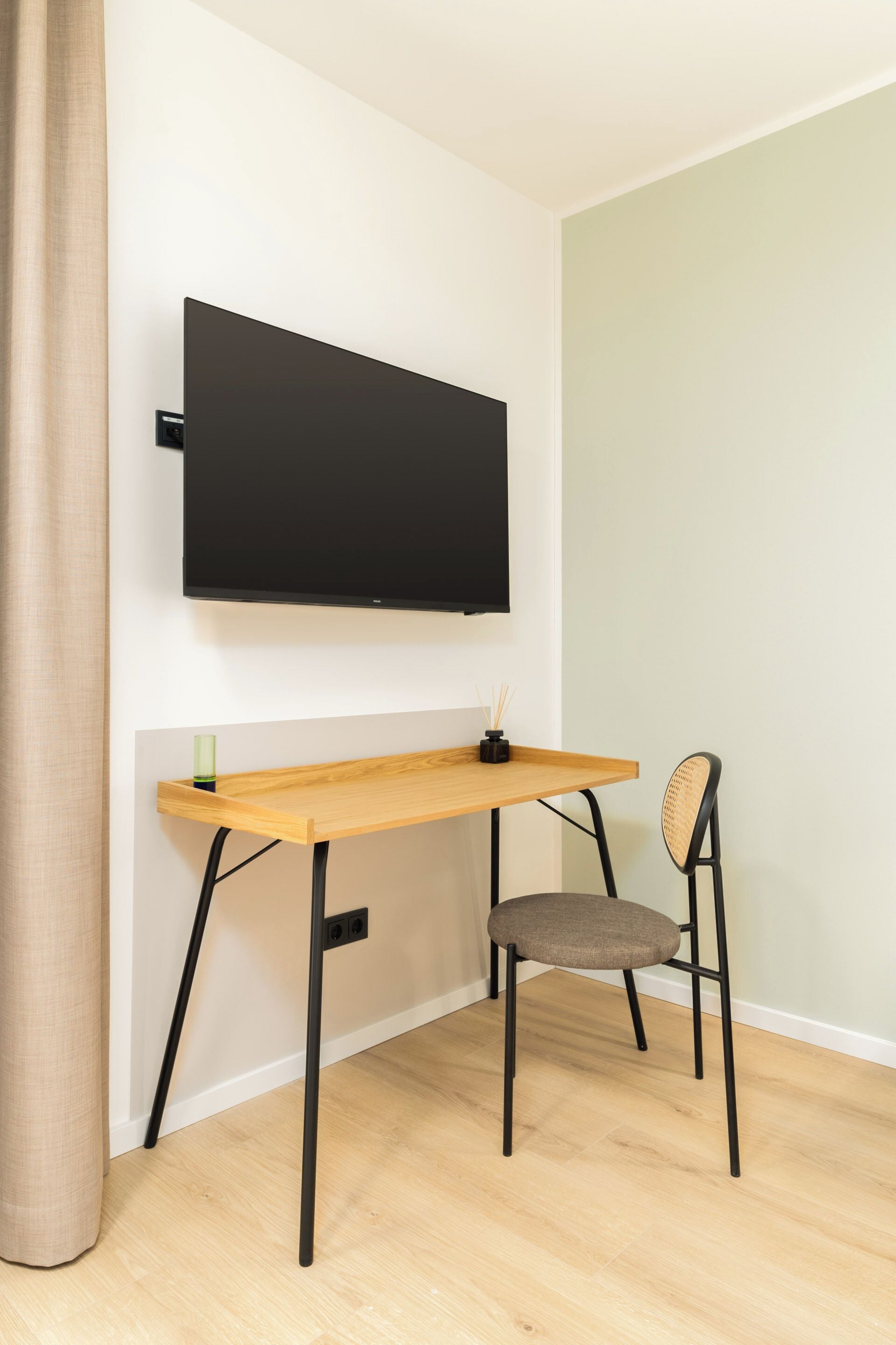 A room with a wooden desk, a chair, a television mounted on the wall, and a curtain.