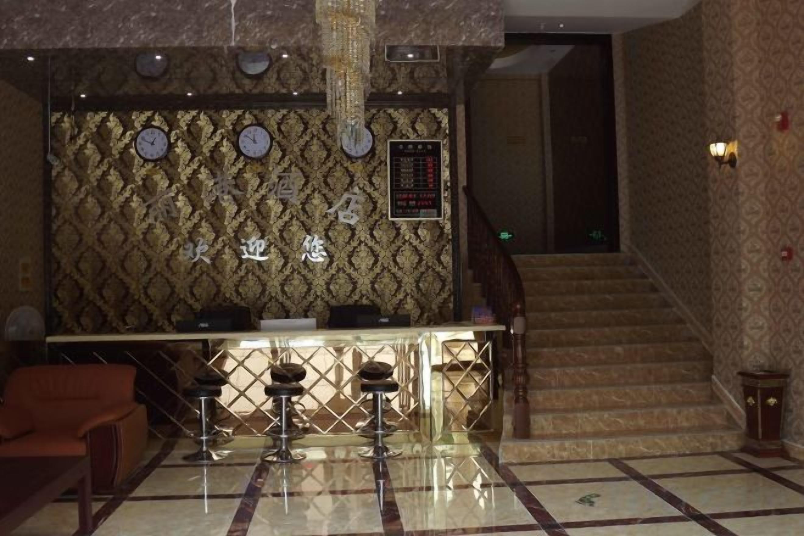 A hotel lobby with a reception desk, bar stools, a patterned wall, clocks, and a staircase.