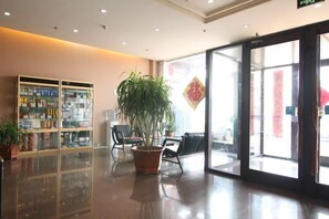 A hotel lobby with a reception desk, a large potted plant, a glass display case, and a large glass door leading outside. - Thank You Junyi Inn Huozhou Gulou (Huozhou)