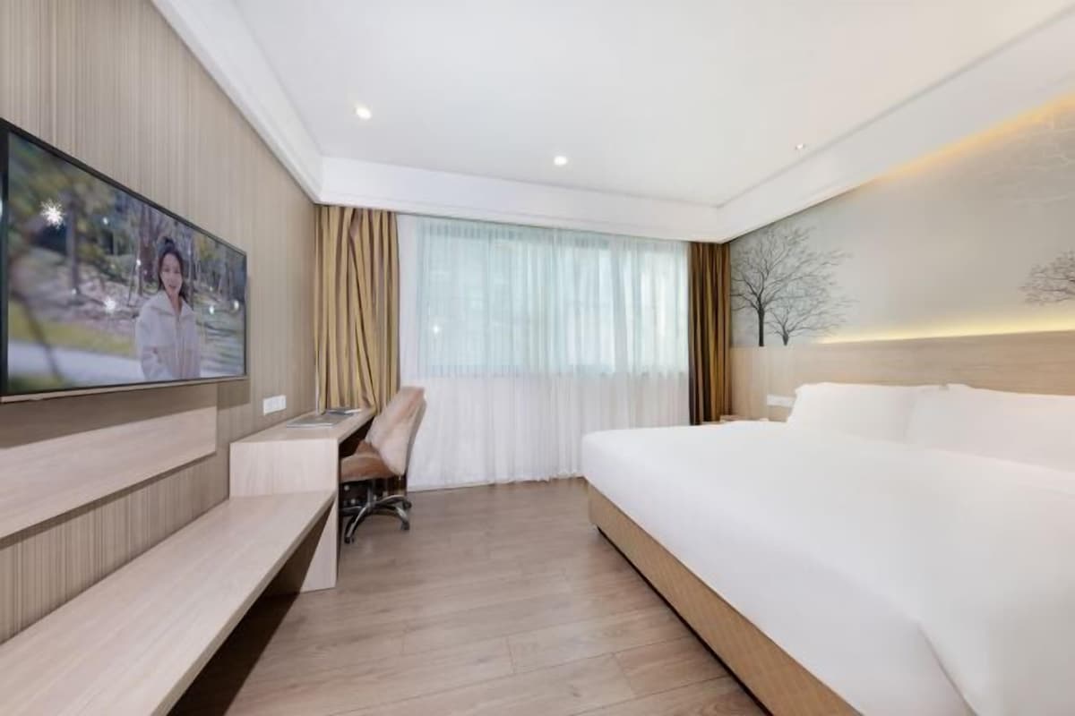 A modern hotel room with a large bed, a desk, a chair, a TV mounted on the wall, and a large window with curtains.