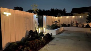 Spazio esterno ben illuminato con recinzione in legno, vialetto in cemento e diverse piante e arbusti.