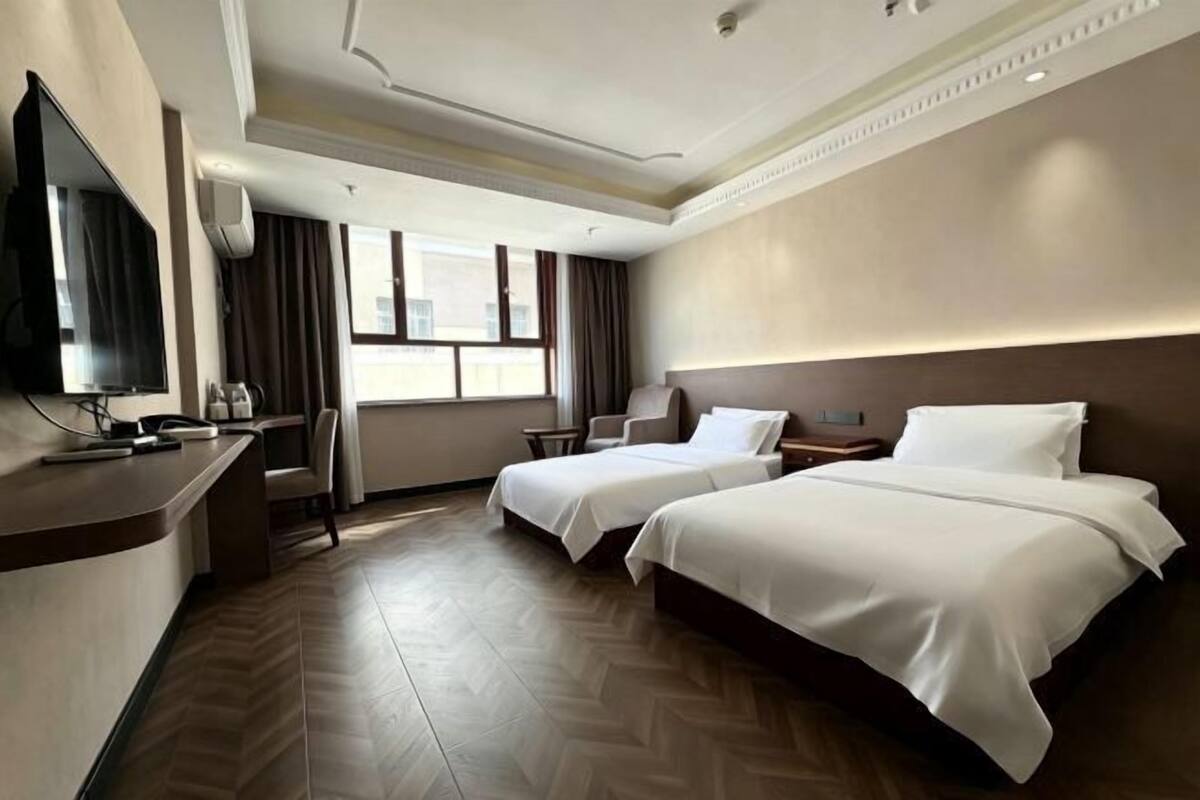 A hotel room with two beds, a desk with a flat-screen TV, a chair, a window with curtains, and a herringbone-patterned floor.