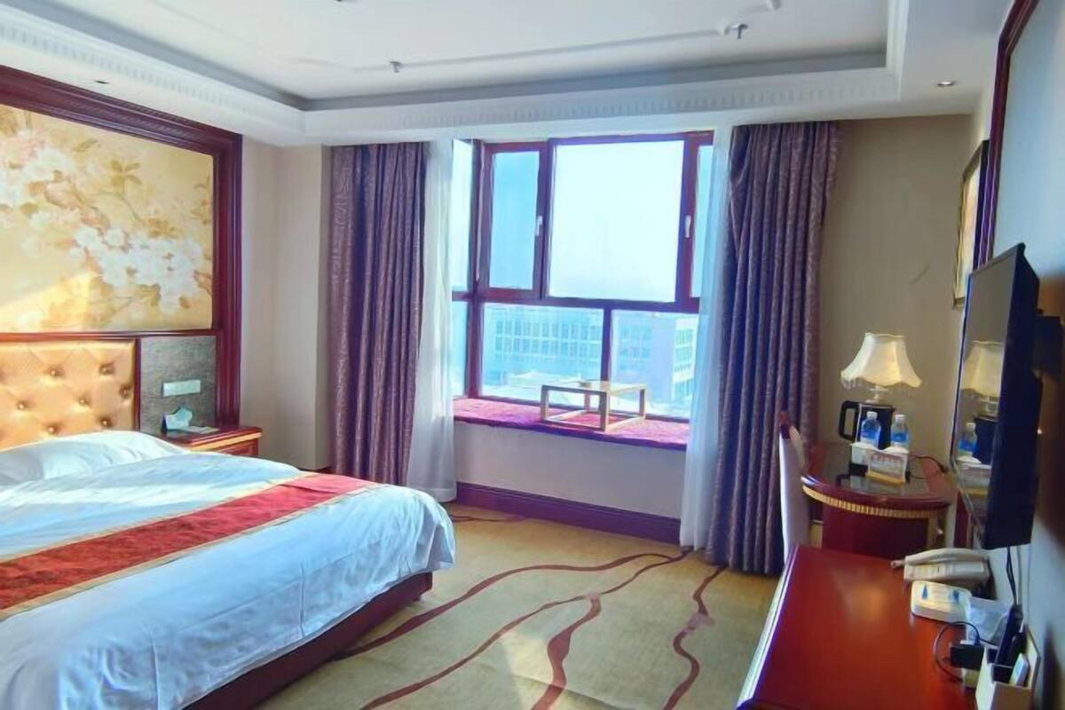 A hotel room with a large bed, a desk, a chair, a window with a view, and a painting on the wall.