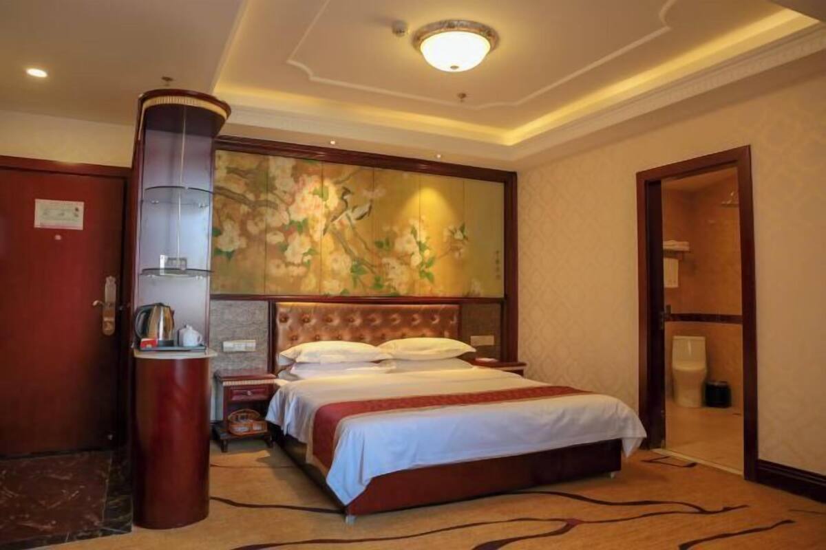 A hotel room with a large bed, a wooden headboard, a wall-mounted artwork, a bedside table with a lamp, and a door leading to a bathroom.