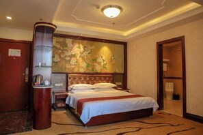 A hotel room with a large bed, a wooden headboard, a wall-mounted artwork, a bedside table with a lamp, and a door leading to a bathroom.
