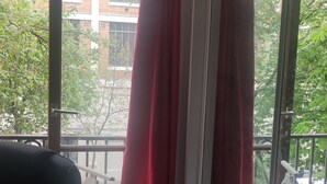 Ein Zimmer mit großem Fenster, roten Vorhängen, einem schwarzen Bürostuhl und einer weißen Heizung.