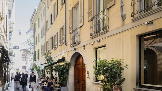 A narrow street with buildings on either side, outdoor seating areas, and a large arched entrance. - Easylife - Delicious Studio in Brera/Duomo (Milano)