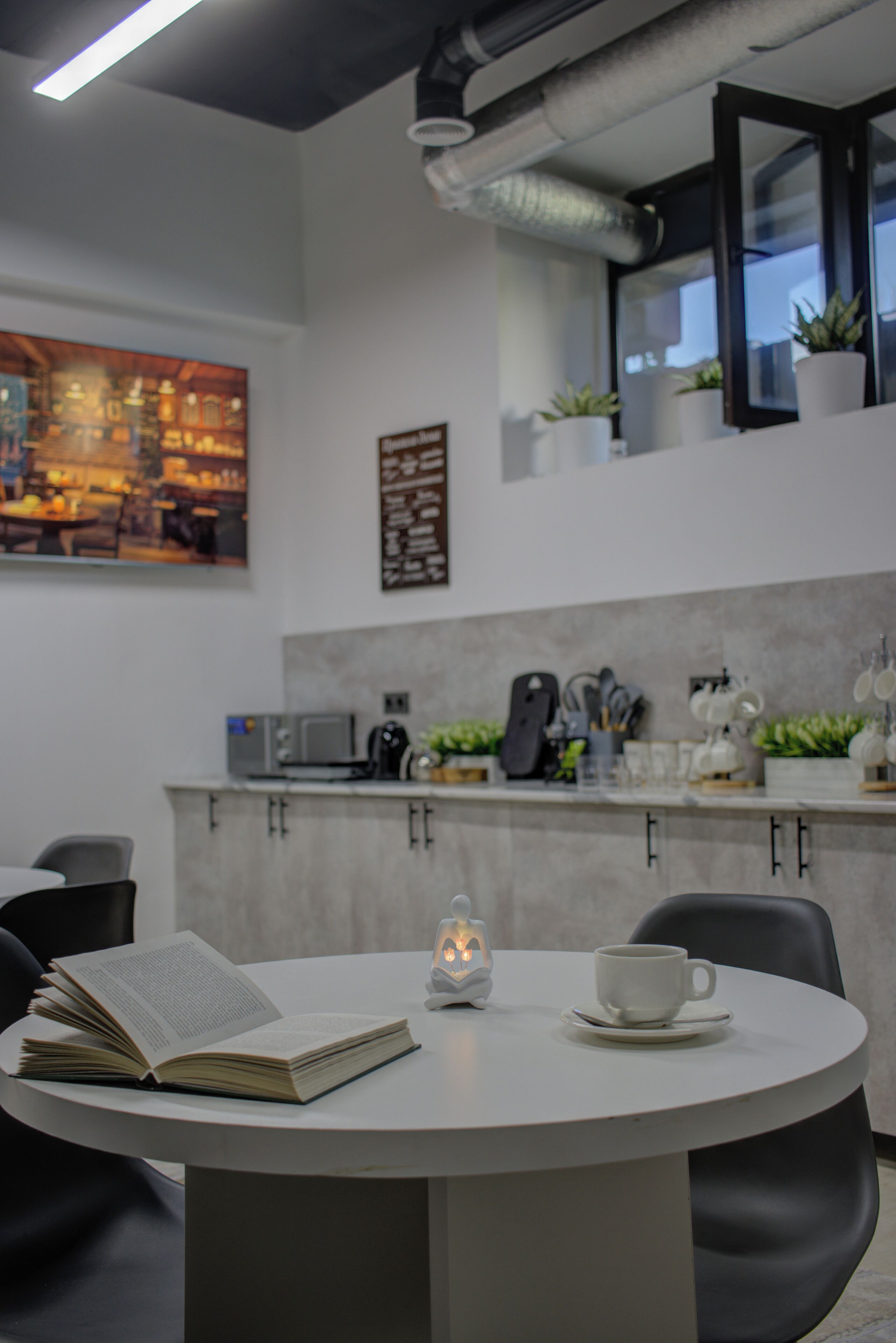 A modern kitchen with a dining table, chairs, and a book. There are plants on the windowsill and a painting on the wall.
