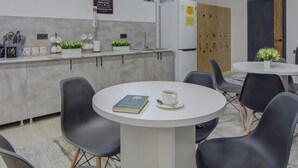 A modern kitchen with a white countertop, black chairs, and a round table. There are cabinets, a refrigerator, and a window with plants. - Hostel Pro (Bishkek)