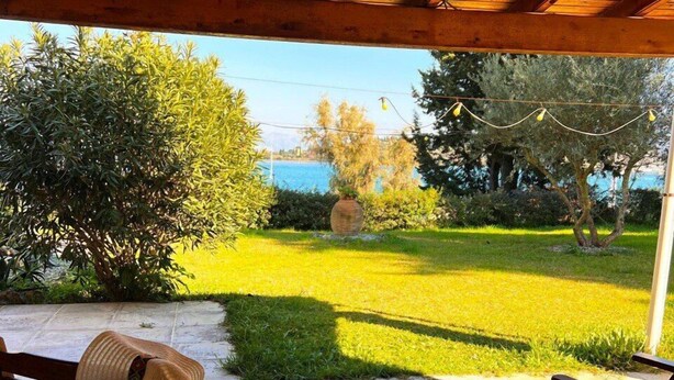 A covered patio with wooden furniture, a table with a bowl of fruit, and a view of a grassy area and trees. - SV Apartment with sea view (Corfu)