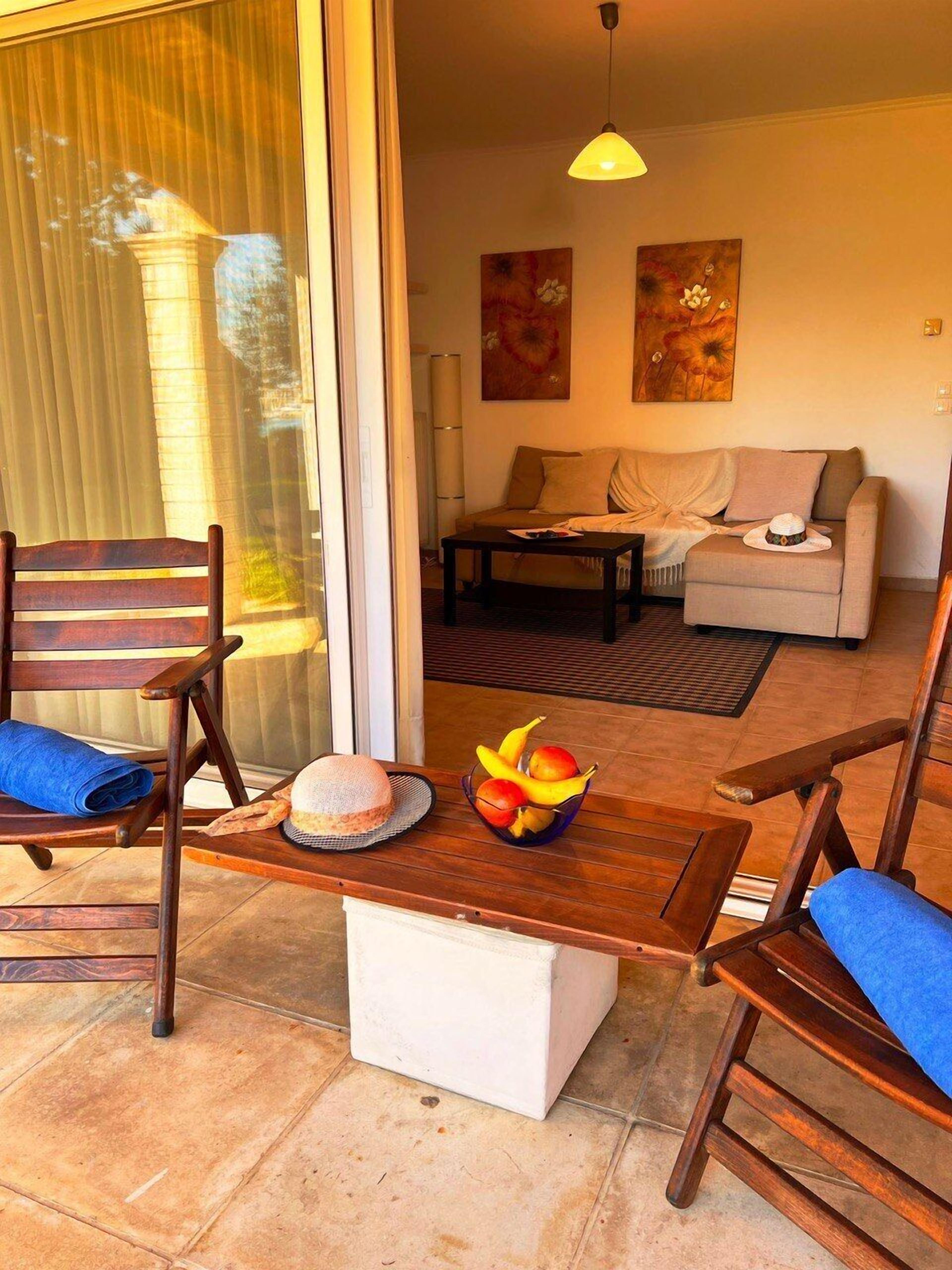 A cozy living area with a sofa, a coffee table, and a dining table with chairs. There is a window with blinds, a painting on the wall, and a fruit bowl on the table.
