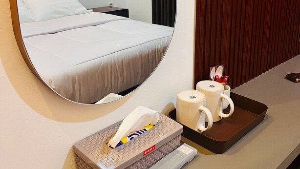 Una habitación de hotel con cama, una mesita de noche con caja de pañuelos, una bandeja con dos tazas y un espejo que refleja la cama y una pared con paneles de madera verticales.