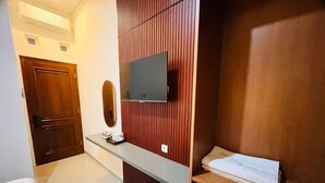 Une chambre d’hôtel avec une porte en bois, un téléviseur à écran plat fixé sur un panneau mural en bois, un petit bureau avec une tasse et une assiette, une chaise grise et une étagère avec une serviette pliée.