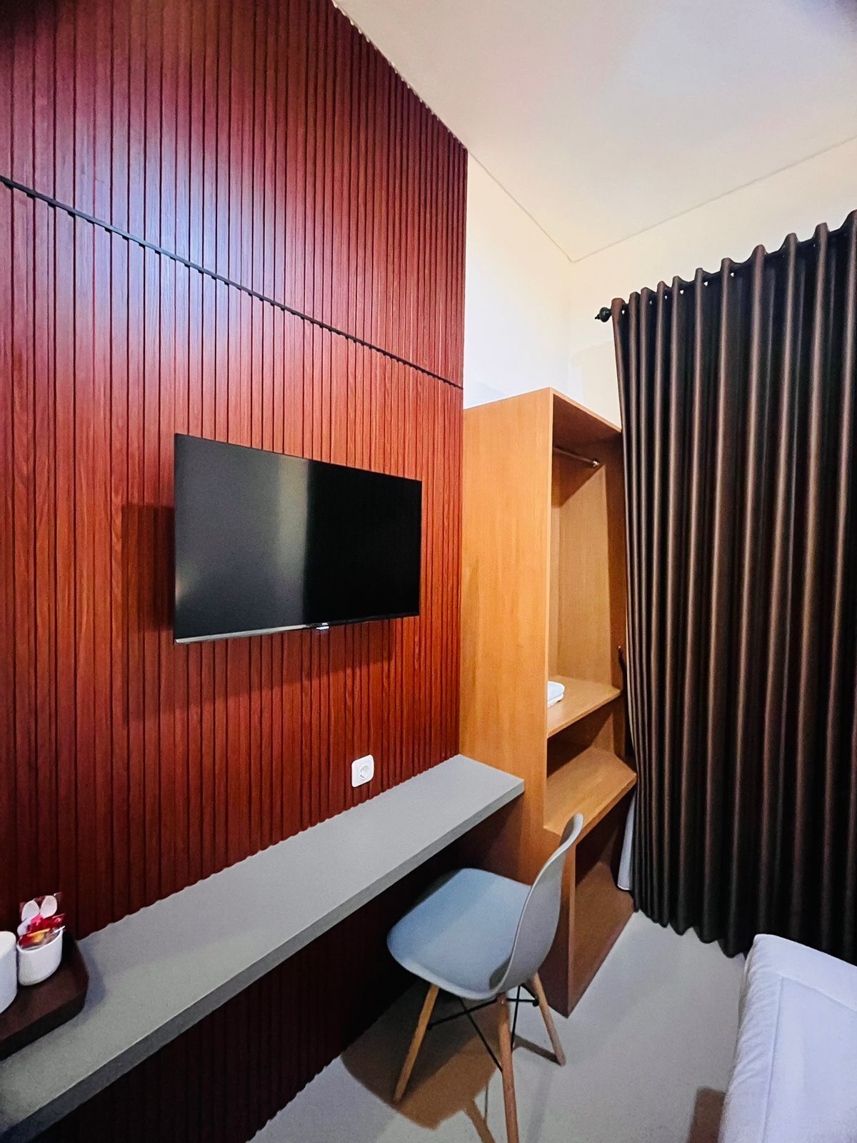 A hotel room with a TV mounted on a wooden wall, a desk with a chair, a shelf, and a bed with curtains.