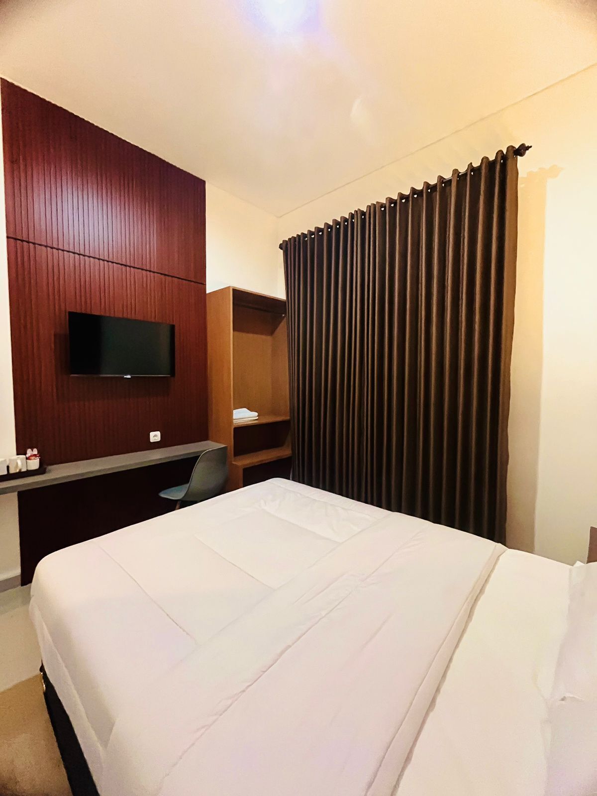 A hotel room with a large bed, a flat-screen TV mounted on a wooden panel wall, a desk with a chair, and a window with dark curtains.