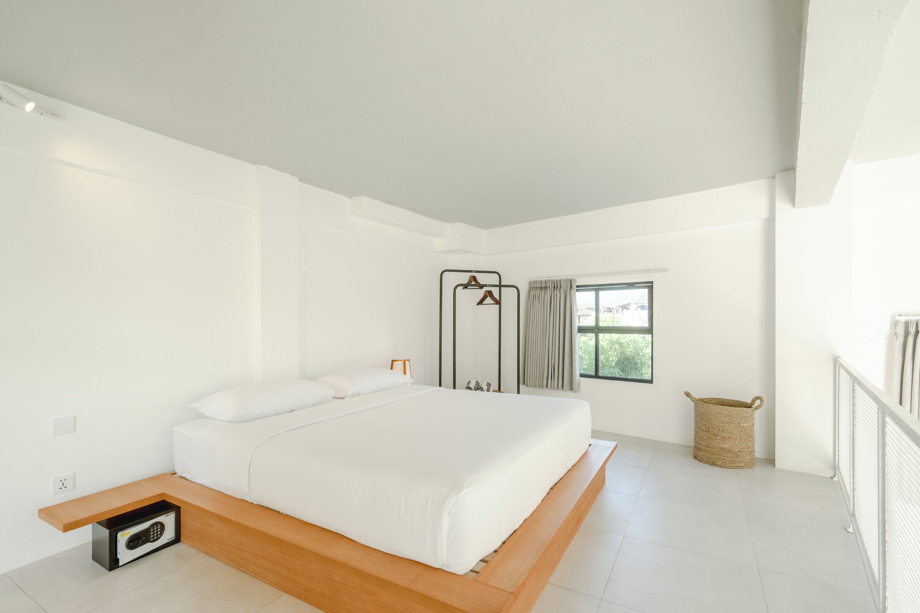 A modern bedroom with a large bed, a small bedside table, a window with curtains, and a wall-mounted radiator.