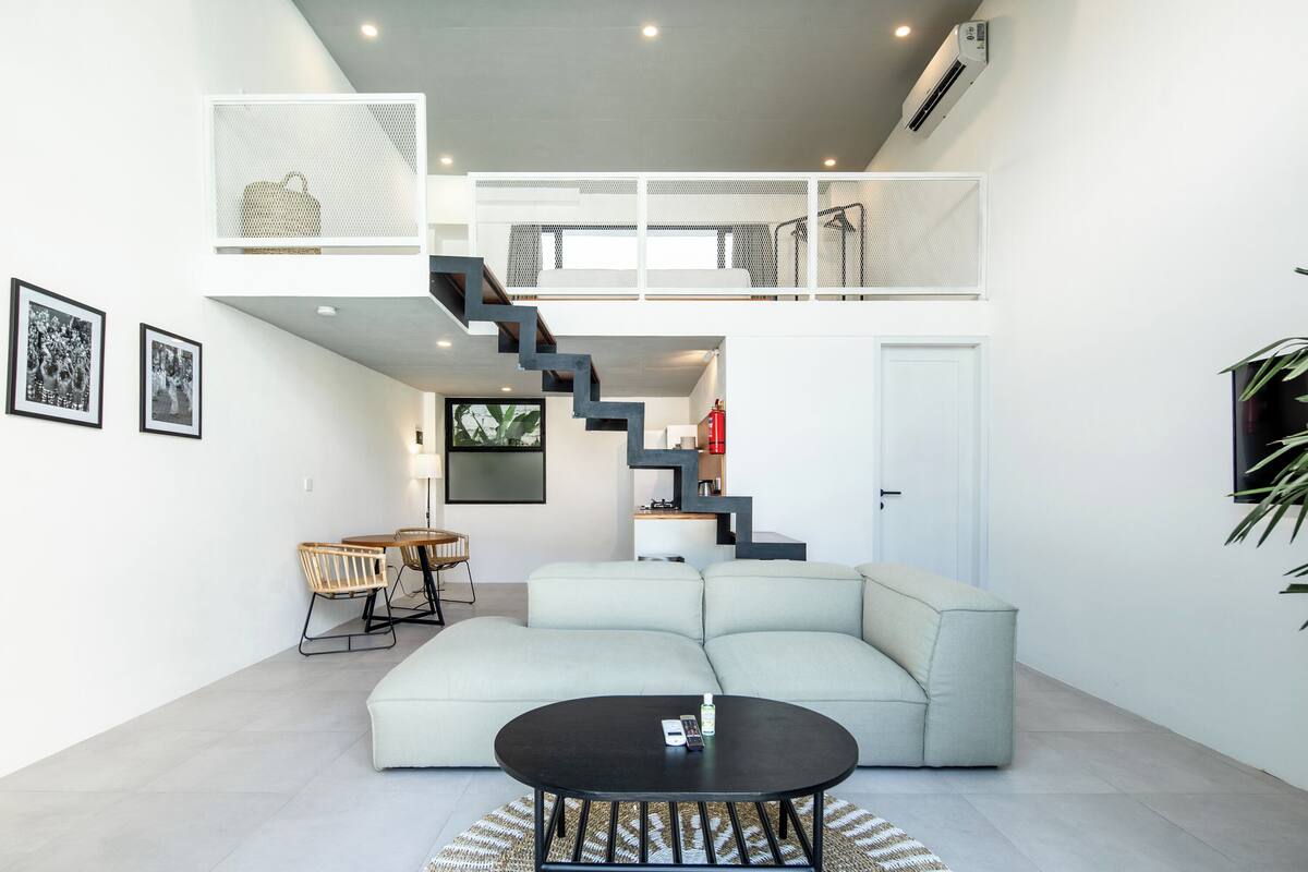 Een moderne woonkamer met een lichtgekleurde modulaire bank, een ronde salontafel en een trap naar een hoger gelegen verdieping.