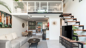 A modern living room with a sofa, coffee table, and a TV. There is a staircase leading to an upper level with a balcony.