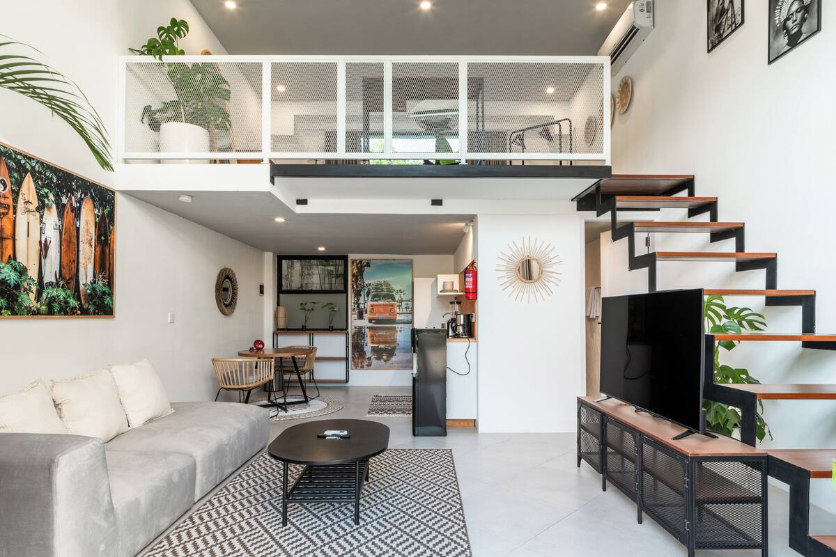 Een moderne woonkamer met een bank, salontafel en een televisie. Er is een trap die leidt naar een hoger niveau met een balkon.